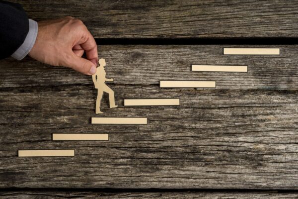 Businessman climbing the steps to success in a conceptual image with paper silhouette cutouts of a man and a human hand helping him climb over rustic wood with copy space.